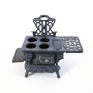 May include: A black cast iron miniature stove featuring four circular burner openings on the top surface, a decorative back splash, and side shelves. The front door is embossed with the text CRESCENT and a star symbol, supported by four legs.