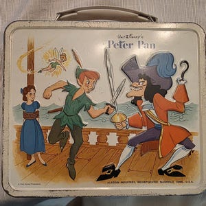 May include: Vintage Peter Pan lunchbox featuring Peter Pan, Captain Hook, Tinkerbell, and Wendy. The lunchbox has a white background with a silver handle and a colorful illustration of the characters in a battle scene. The text "Walt Disney's Peter Pan" is visible.