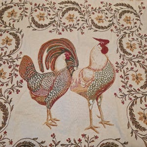 May include: A woven fabric textile featuring two roosters standing side by side in the center. The roosters are rendered in shades of red, green, brown, and cream. A decorative border of scrolling vines, leaves, and small red flowers frames the birds.