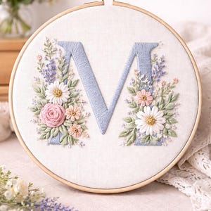 May include: Embroidered floral letter "M" in a wooden hoop. The light blue letter is surrounded by pink, white, and peach flowers with green leaves. The embroidery is on a white fabric background.