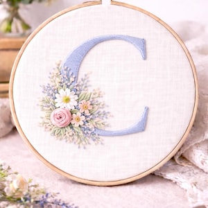 Può includere: Ricamo della lettera "C" in azzurro, circondata da una composizione floreale di fiori rosa, bianchi e verde chiaro, all'interno di un telaio di legno. Lo sfondo è un tessuto morbido bianco sporco.