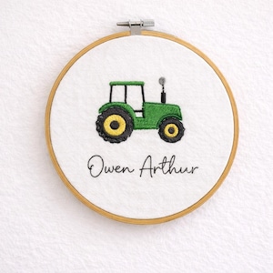May include: Embroidered wall art showing a green tractor with black and yellow wheels on a white background. The name "Owen Arthur" is written in cursive below the tractor. The design is framed in a wooden embroidery hoop.