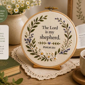 Pode incluir: Arte de bordado em bastidor com o texto "The Lord is my shepherd" e Salmo 23:1. O design apresenta elementos florais em branco, verde e roxo. Um vaso de margaridas, um livro e uma xícara de café também estão na imagem.