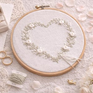Puede incluir: Un bastidor de bordado con un diseño floral en forma de corazón, un anillo en una caja beige y dos alianzas de boda doradas. Un carrete de hilo y un ramo de rosas blancas también están presentes, creando una escena romántica con temática de boda.