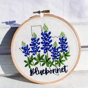 May include: Embroidered hoop art depicting bluebonnet flowers and the outline of Texas. The flowers are a vibrant blue with green leaves and buds. The word "Bluebonnet" is written in cursive below the flowers.