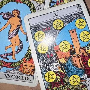 May include: Two tarot cards are displayed. One card features a nude figure with a white sash, surrounded by a blue background and the word "WORLD." The other card shows a scene with figures, a tower, and yellow pentacles.