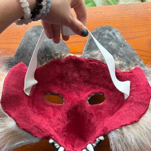 May include: A red and white furry mask with eye holes and a toothy grin. The mask has gray ears and white elastic straps. The mask is held up by a hand with bracelets.