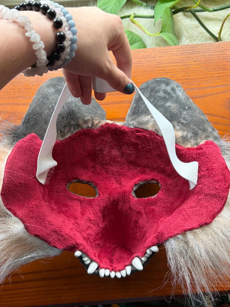 May include: A red and white furry mask with eye holes and a toothy grin. The mask has gray ears and white elastic straps. The mask is held up by a hand with bracelets.