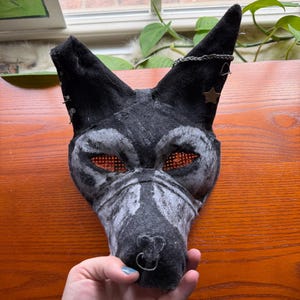 May include: A handmade black and gray wolf mask with orange mesh eyes, a silver nose ring, and a star-shaped embellishment. The mask is made of felt and has a textured appearance, suitable for cosplay or costume events.
