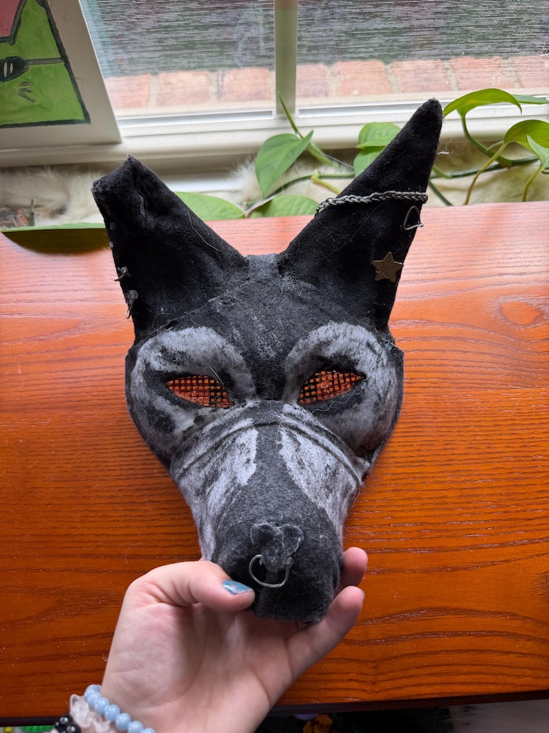 May include: A handmade black and gray wolf mask with orange mesh eyes, a silver nose ring, and a star-shaped embellishment. The mask is made of felt and has a textured appearance, suitable for cosplay or costume events.