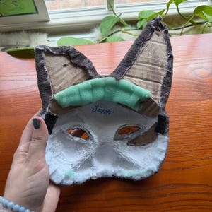 May include: A handmade cat mask with brown ears, a white face, and eye cutouts. The mask has a green foam piece across the forehead and black elastic straps. The name "Jax" is written on the mask.