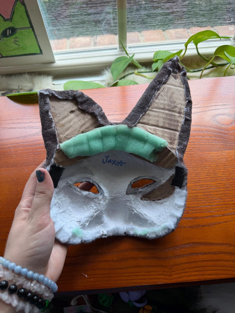 May include: A handmade cat mask with brown ears, a white face, and eye cutouts. The mask has a green foam piece across the forehead and black elastic straps. The name "Jax" is written on the mask.