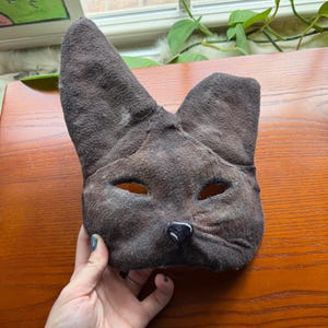 May include: A brown cat mask with eye holes and a black nose. The mask is made of a textured material and has large, pointed ears. The mask is held up by a hand.