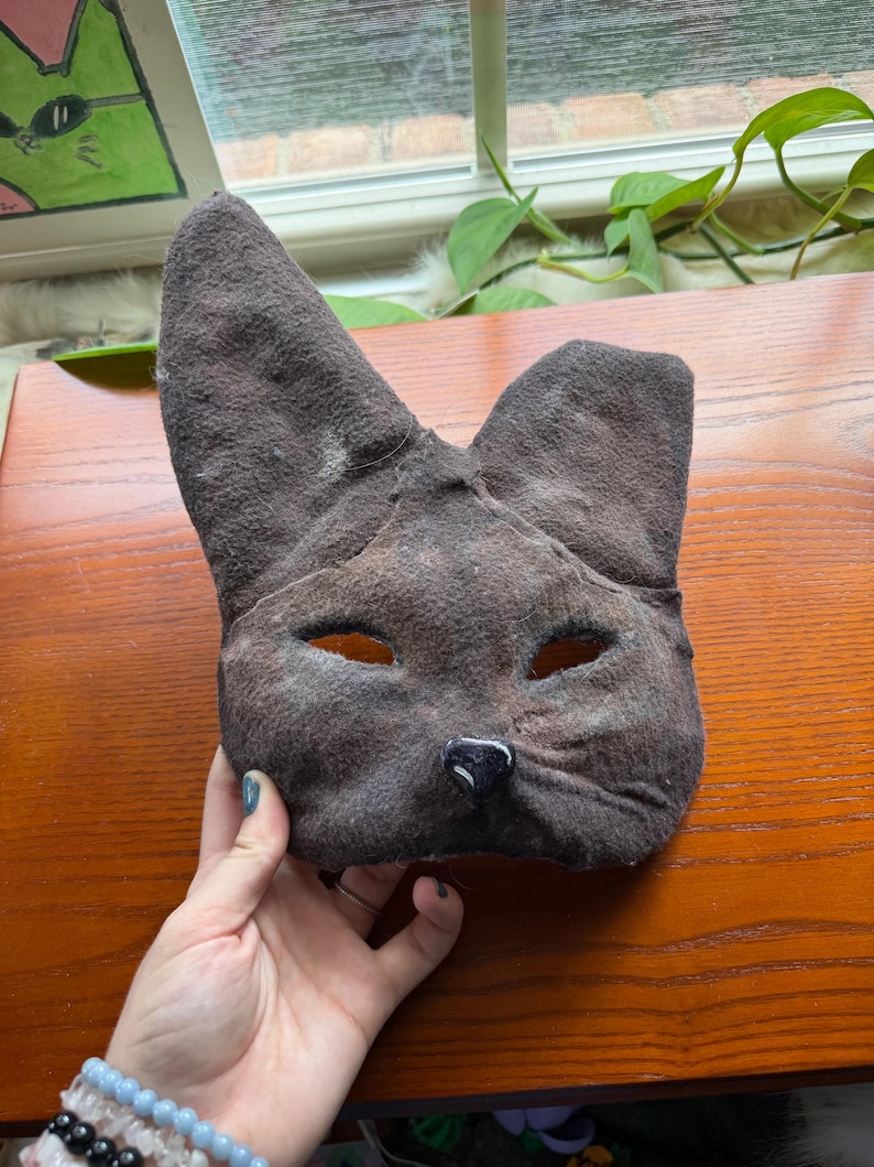 May include: A brown cat mask with eye holes and a black nose. The mask is made of a textured material and has large, pointed ears. The mask is held up by a hand.