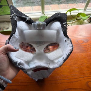 May include: A handmade, black and white cat mask with mesh eye coverings and foam padding. The mask has a red mouth detail and is designed to be worn. The interior is white, and the exterior is black.