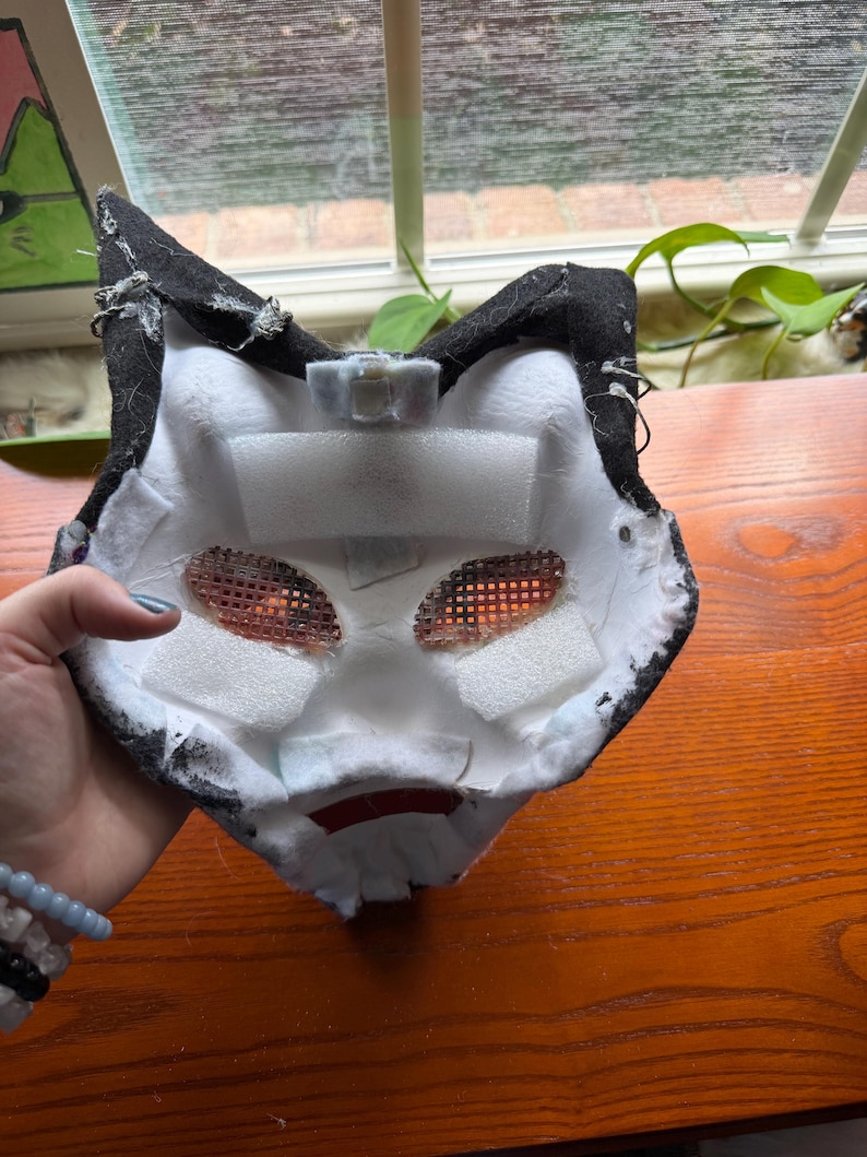 May include: A handmade, black and white cat mask with mesh eye coverings and foam padding. The mask has a red mouth detail and is designed to be worn. The interior is white, and the exterior is black.