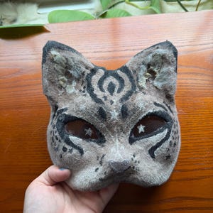 May include: A handmade cat mask with a spotted pattern in shades of gray and black. The mask features mesh eye coverings and is designed to resemble a feline face. The mask is made of felt and is suitable for costume or cosplay.