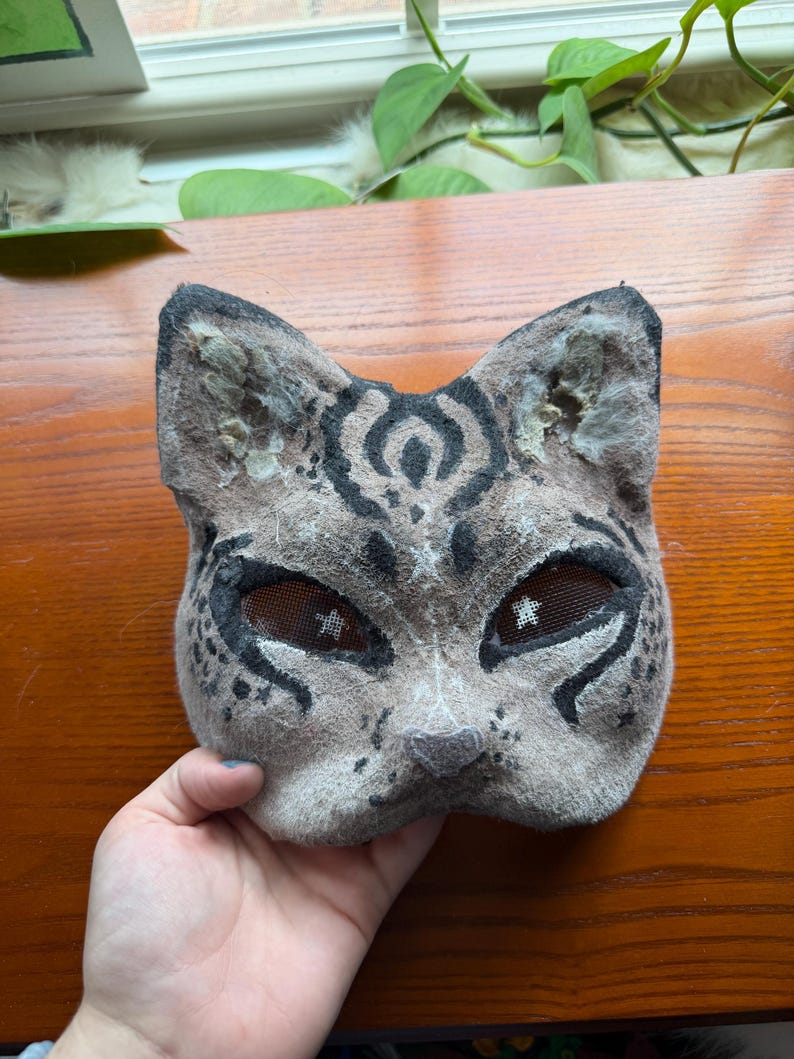 May include: A handmade cat mask with a spotted pattern in shades of gray and black. The mask features mesh eye coverings and is designed to resemble a feline face. The mask is made of felt and is suitable for costume or cosplay.