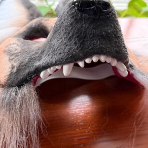 May include: Close-up of a wolf mask with a black nose, gray fur, and white teeth. The mask has a red interior and is made of a soft, textured material. The mask is designed to be worn.