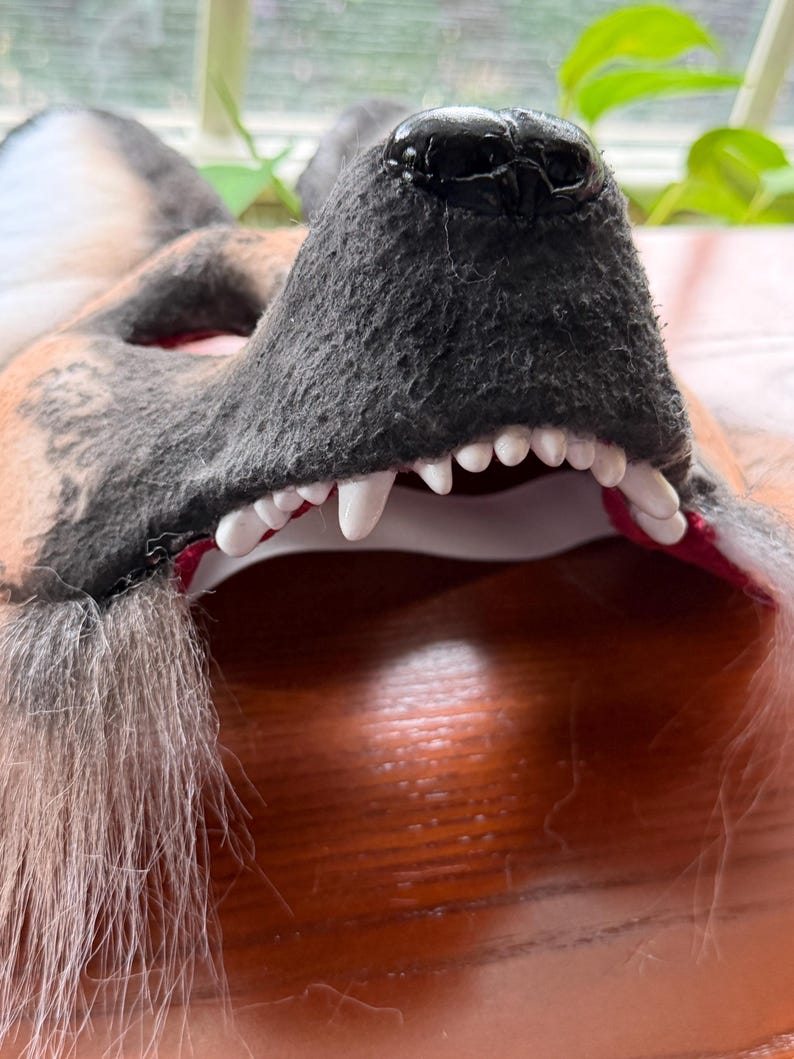 May include: Close-up of a wolf mask with a black nose, gray fur, and white teeth. The mask has a red interior and is made of a soft, textured material. The mask is designed to be worn.