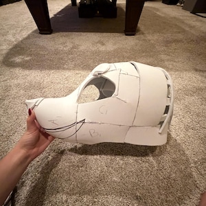 May include: A white foam animal mask base featuring a snout, large eye openings, and rounded ear structures. The surface is constructed from multiple joined panels with visible black ink markings, including the letters A, B, and C, and a drawn arrow.