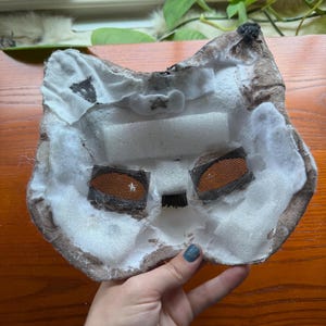 May include: A handmade cat mask with a brown and white color scheme. The mask features mesh eye coverings and is constructed with foam and fabric. The interior is white, and the edges are brown.
