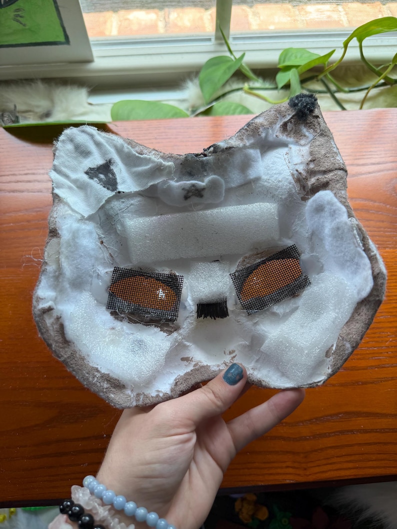 May include: A handmade cat mask with a brown and white color scheme. The mask features mesh eye coverings and is constructed with foam and fabric. The interior is white, and the edges are brown.