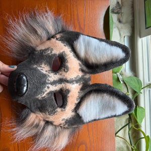 May include: A handmade dog mask with a black nose, tan and black fur pattern, and white-lined ears. The mask has eye openings and is made of soft, plush materials. The mask is designed for costume or cosplay use.