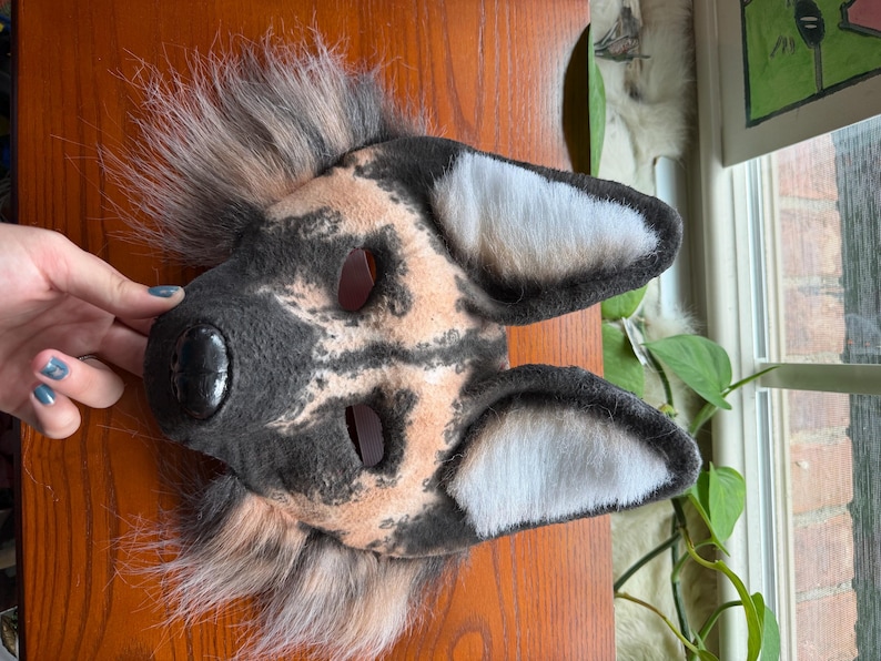 May include: A handmade dog mask with a black nose, tan and black fur pattern, and white-lined ears. The mask has eye openings and is made of soft, plush materials. The mask is designed for costume or cosplay use.