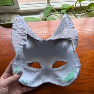 May include: A handmade, white and gray cat mask with eye cutouts. The mask has a textured, felt-like exterior and a soft, white interior. Green accents are visible near the edges. A small metal clasp is attached to the back for hanging.