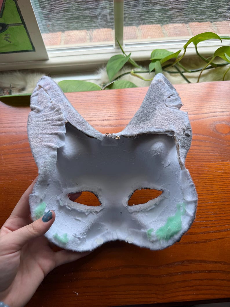 May include: A handmade, white and gray cat mask with eye cutouts. The mask has a textured, felt-like exterior and a soft, white interior. Green accents are visible near the edges. A small metal clasp is attached to the back for hanging.