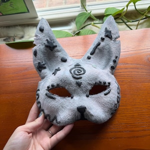May include: A gray cat mask with black accents. The mask has eye holes, a nose, and pointed ears. Black dots and star shapes decorate the ears. A spiral design is centered on the forehead. The mask is handmade.