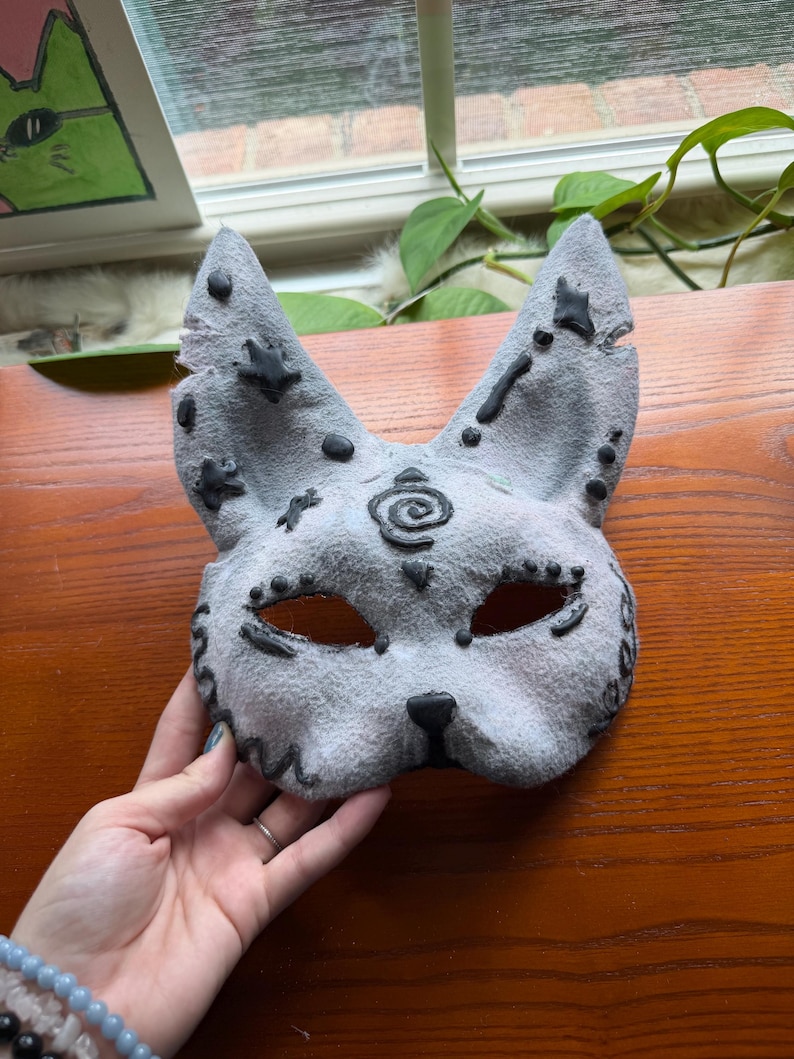 May include: A gray cat mask with black accents. The mask has eye holes, a nose, and pointed ears. Black dots and star shapes decorate the ears. A spiral design is centered on the forehead. The mask is handmade.