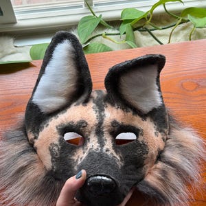May include: A handmade African wild dog mask with a black, tan, and white color scheme. The mask features large ears, eye holes, and a detailed snout. The mask is made of soft, plush materials and has a fur trim.