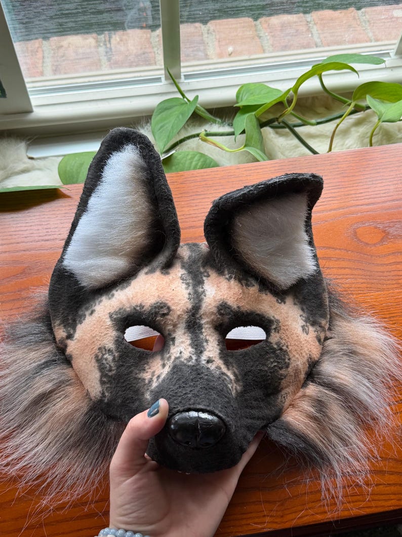May include: A handmade African wild dog mask with a black, tan, and white color scheme. The mask features large ears, eye holes, and a detailed snout. The mask is made of soft, plush materials and has a fur trim.