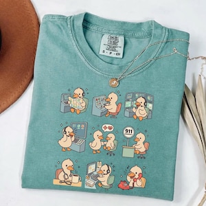 Puede incluir: Una camiseta Comfort Colors de color turquesa con patos de dibujos animados en diversas situaciones de servicios de emergencia. Los patos se representan usando computadoras, teléfonos y equipos médicos. La camiseta también tiene el logotipo de Comfort Colors e información de tallas.