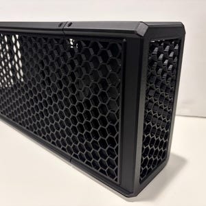 May include: A black rectangular metal enclosure with a honeycomb mesh design. The enclosure has a solid frame and is designed with a geometric pattern. The object is likely a component for electronics or a similar application.