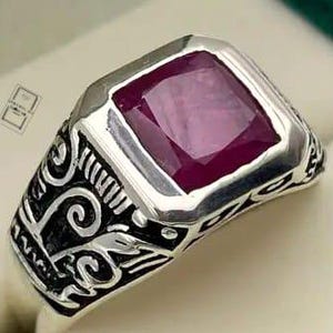 Peut inclure: Une bague en argent ornée d'une grande pierre précieuse rouge foncé de taille carrée au centre. L'anneau est fabriqué en argent poli avec des motifs de volutes gravés sombres et complexes sur les côtés, offrant une finition décorative contrastée.