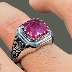 May include: A silver ring featuring a large, faceted, cushion-cut deep pinkish-red gemstone set in a prong setting. The band is crafted from polished silver with intricate, dark, engraved scrollwork patterns along the sides of the ring shank.