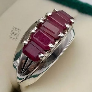 May include: A silver ring featuring three rectangular, emerald-cut deep red rubies set in a row. The metal band has a polished, reflective finish with a smooth, curved design. A small, square logo is visible on the side of the ring setting.