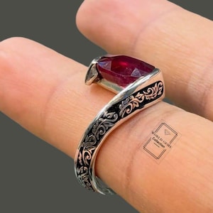 May include: A silver ring featuring an elongated, marquise-cut deep red gemstone set in a band with intricate, dark-oxidised scrollwork engravings. The metal band displays a detailed floral-inspired pattern across its entire visible surface.
