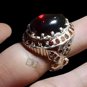 Puede incluir: Un anillo de plata con una gran piedra preciosa cabujón ovalada de color rojo oscuro en el centro, rodeada por un halo de piedras redondas más pequeñas de color rojo oscuro. El aro está decorado con pequeñas piedras rojas y el texto World Gems Collection.