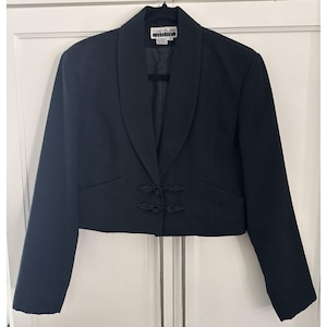 May include: A cropped black blazer featuring a shawl collar, long sleeves, and two decorative frog closures at the waist. The interior label displays the text Contempo Casuals. The garment is constructed with a solid black fabric throughout.