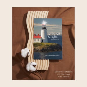 May include: A softcover notebook with a matte finish featuring a lighthouse image on the cover. The text reads Be the Salt & Light... Matthew 5:13-16 and Daily Reflections Notebook. It includes 100 lined pages and is displayed with a wavy wooden stand.