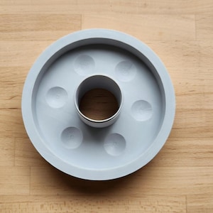 Puede incluir: Un posavasos de hormigón gris claro con un anillo central de metal y seis hendiduras circulares. El posavasos circular se encuentra sobre una superficie de madera marrón claro. Diseño minimalista y moderno.