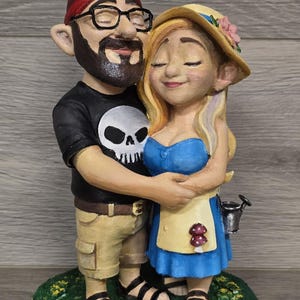 Puede incluir: Una figura caprichosa de una pareja abrazándose, diseñada como gnomos de jardín. El gnomo masculino lleva una camisa negra con una calavera, gafas y un gorro rojo. La gnomo femenina lleva un vestido azul y un sombrero de paja. Se paran sobre una base verde.