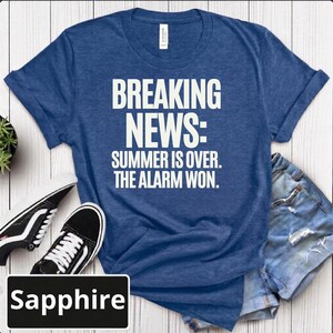 May include: A heathered sapphire blue short-sleeved t-shirt with white text that reads: BREAKING NEWS: SUMMER IS OVER. THE ALARM WON. The shirt features a crew neckline and is displayed alongside black and white trainers and denim shorts.