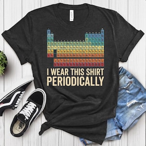 May include: A dark grey short-sleeved t-shirt featuring a colourful periodic table of elements graphic centred on the chest. Below the table, the text reads I WEAR THIS SHIRT PERIODICALLY in large, bold, light-coloured capital letters.