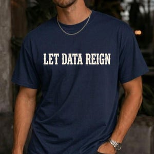 以下が含まれることがあります： 胸元に白の掠れたフォントで「LET DATA REIGN」とプリントされた、ネイビーブルーの半袖Tシャツ。この衣類はクラシックなクルーネックで、シルバーのチェーンネックレスとシルバーの腕時計を合わせて着用されています。