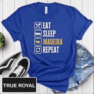 Puede incluir: Una camiseta de manga corta en azul real con un diseño gráfico blanco de cuatro cuadrados apilados que contienen iconos de comer, dormir, la isla de Madeira y repetir, con el texto EAT SLEEP MADEIRA REPEAT en blanco y dorado.